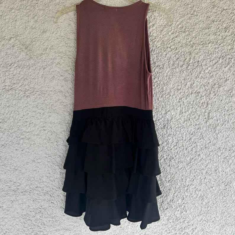 Puede incluir: Vestido sin mangas con un corpi&ntilde;o malva y una falda negra con volantes en capas. El vestido est&aacute; colgado de una percha de pl&aacute;stico transparente sobre un fondo texturizado blanquecino. El vestido parece estar hecho de tela suave.