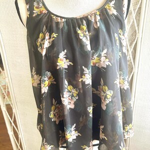 K&ouml;nnte beinhalten: Eine &auml;rmellose, dunkelgraue Bluse mit floralem Muster. Die Blumen sind in Gelb-, Rosa- und Blaut&ouml;nen gehalten. Die Bluse hat einen Rundhalsausschnitt und ist aus einem transparenten Stoff gefertigt.