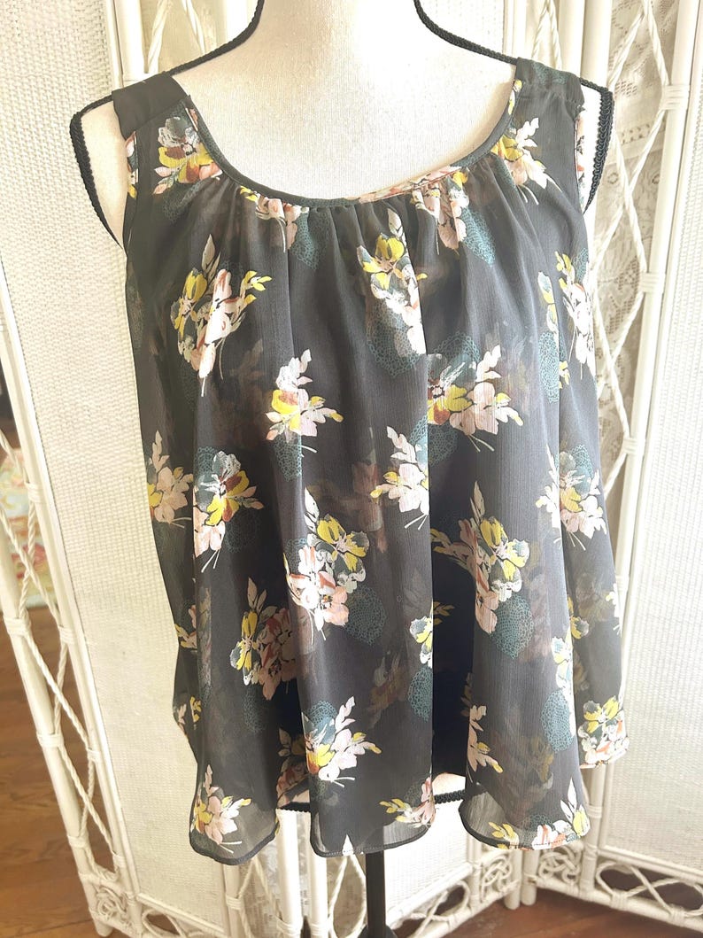 K&ouml;nnte beinhalten: Eine &auml;rmellose, dunkelgraue Bluse mit floralem Muster. Die Blumen sind in Gelb-, Rosa- und Blaut&ouml;nen gehalten. Die Bluse hat einen Rundhalsausschnitt und ist aus einem transparenten Stoff gefertigt.