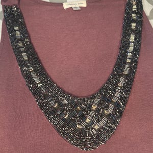 Puede incluir: Camiseta sin mangas de color malva con un dise&ntilde;o de collar de cuentas. El collar presenta una forma de cuello en V con cuentas de color gris oscuro y plateado. La parte superior tiene un escote redondo y la etiqueta dice "silence + noise".