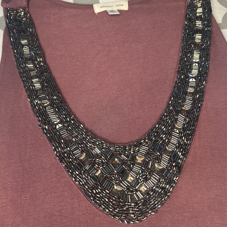 Puede incluir: Camiseta sin mangas de color malva con un dise&ntilde;o de collar de cuentas. El collar presenta una forma de cuello en V con cuentas de color gris oscuro y plateado. La parte superior tiene un escote redondo y la etiqueta dice "silence + noise".
