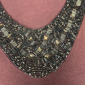 Puede incluir: Primer plano de una blusa color malva con cuello de cuentas. El cuello en forma de V est&aacute; profusamente adornado con cuentas grises oscuras y plateadas. Las cuentas est&aacute;n dispuestas en un patr&oacute;n geom&eacute;trico.