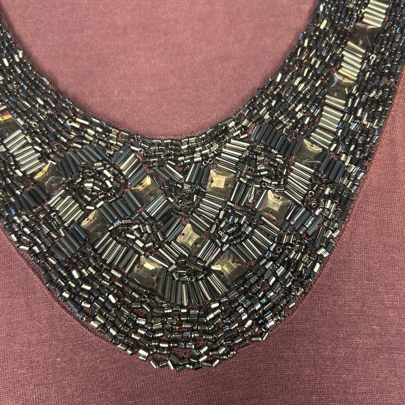 Puede incluir: Primer plano de una blusa color malva con cuello de cuentas. El cuello en forma de V est&aacute; profusamente adornado con cuentas grises oscuras y plateadas. Las cuentas est&aacute;n dispuestas en un patr&oacute;n geom&eacute;trico.