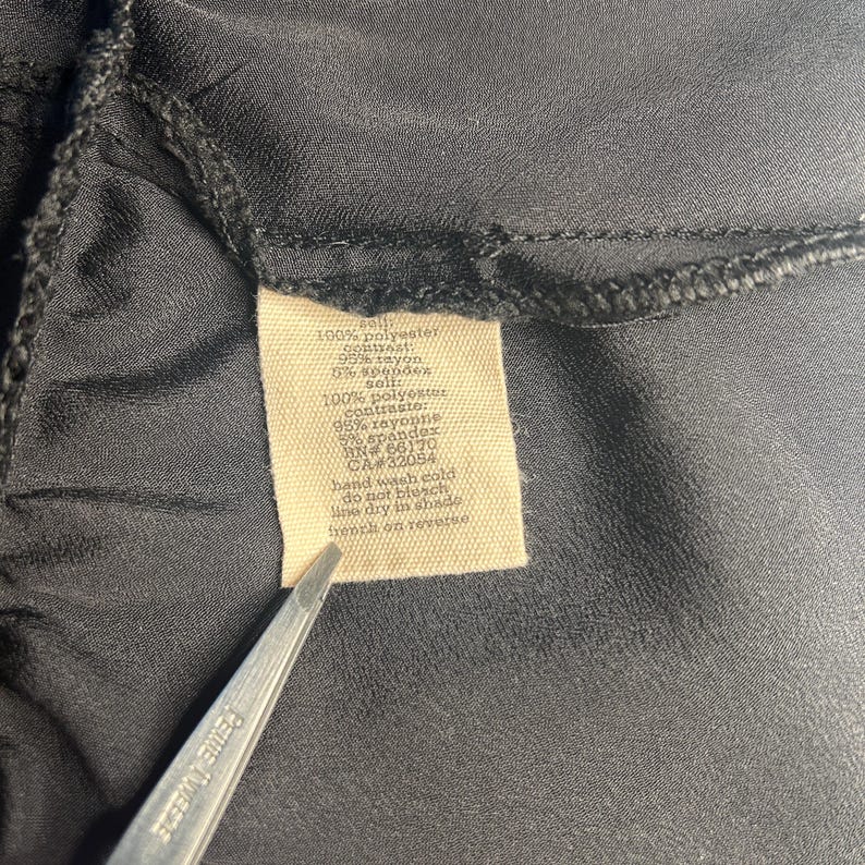 Puede incluir: Primer plano de una tela negra con una costura. Una etiqueta rectangular con texto es visible, junto con unas pinzas. La etiqueta enumera la composici&oacute;n de la tela y las instrucciones de cuidado.