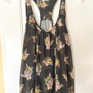 K&ouml;nnte beinhalten: Schwarzes Tanktop mit einem Blumenmuster in Rosa-, Gelb- und Gr&uuml;nt&ouml;nen. Das Tanktop hat ein Racerback-Design und h&auml;ngt an einem durchsichtigen Kunststoffb&uuml;gel.