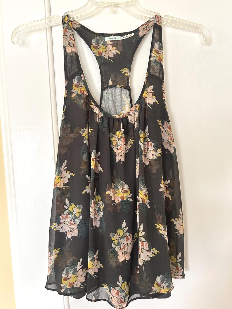 K&ouml;nnte beinhalten: Schwarzes Tanktop mit einem Blumenmuster in Rosa-, Gelb- und Gr&uuml;nt&ouml;nen. Das Tanktop hat ein Racerback-Design und h&auml;ngt an einem durchsichtigen Kunststoffb&uuml;gel.