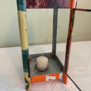 May include: A rectangular metal lantern with glass panels, featuring a white candle inside. The lantern has a mix of colors, including green, orange, and red, with visible text on the sides. The design is vintage and decorative.