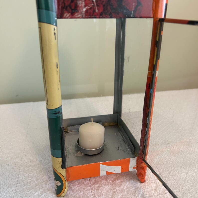 May include: A rectangular metal lantern with glass panels, featuring a white candle inside. The lantern has a mix of colors, including green, orange, and red, with visible text on the sides. The design is vintage and decorative.