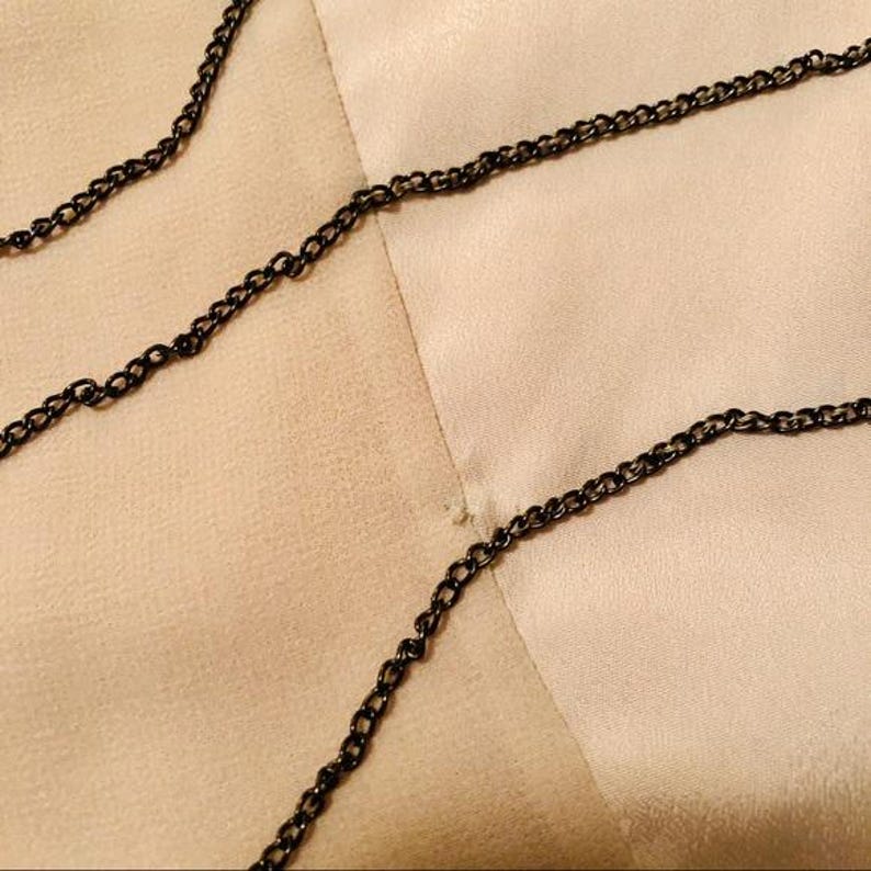 May include: Close-up of a black chain necklace on a light beige fabric background. The chain is made of small, interconnected links. The fabric has a subtle texture and a slight sheen, with a small imperfection visible.