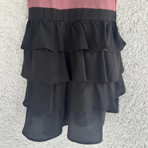 Puede incluir: Vestido sin mangas con corpi&ntilde;o malva y falda negra con volantes escalonados. La falda tiene m&uacute;ltiples capas de tela fluida, creando un aspecto din&aacute;mico y texturizado. El vestido es adecuado para diversas ocasiones.
