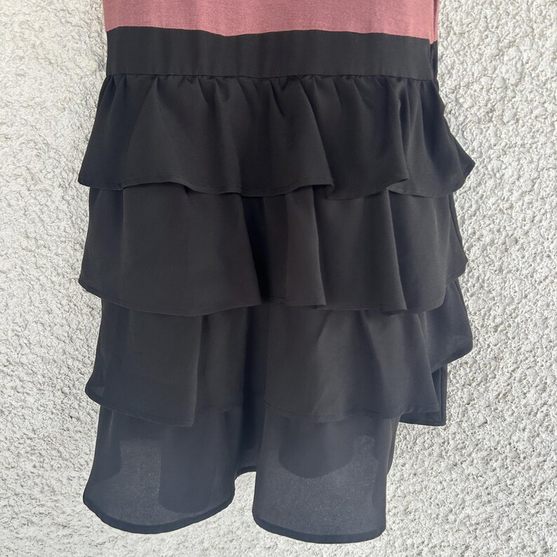 Puede incluir: Vestido sin mangas con corpi&ntilde;o malva y falda negra con volantes escalonados. La falda tiene m&uacute;ltiples capas de tela fluida, creando un aspecto din&aacute;mico y texturizado. El vestido es adecuado para diversas ocasiones.