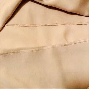 May include: Close-up of a beige fabric with visible stitching. The fabric appears to be smooth and flowing, with a subtle sheen. The stitching is neatly done, adding a touch of detail to the overall design.