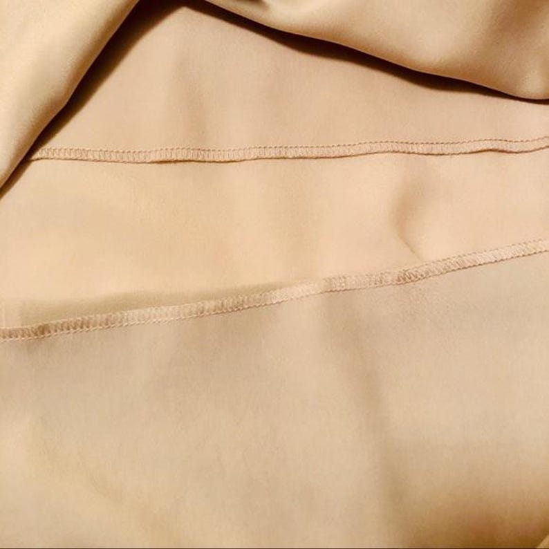 May include: Close-up of a beige fabric with visible stitching. The fabric appears to be smooth and flowing, with a subtle sheen. The stitching is neatly done, adding a touch of detail to the overall design.