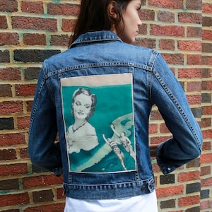 以下が含まれることがあります： 背中にヴィンテージ風のグラフィックがプリントされたデニムジャケット。グラフィックは、女性と水着姿の2人の人物が描かれています。ジャケットはミディアムウォッシュブルーで、下に白いTシャツを着ています。