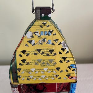 May include: A handmade, upcycled lantern crafted from a repurposed food container. The lantern features a yellow and red color scheme with cut-out patterns, a metal handle, and text from the original packaging. It's a unique, eco-friendly decorative item.