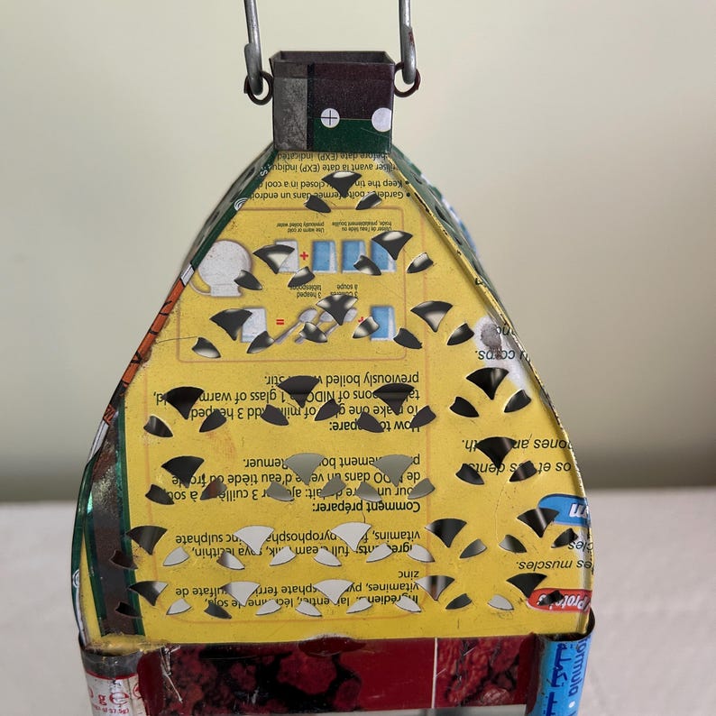 May include: A handmade, upcycled lantern crafted from a repurposed food container. The lantern features a yellow and red color scheme with cut-out patterns, a metal handle, and text from the original packaging. It's a unique, eco-friendly decorative item.