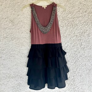 Puede incluir: Vestido sin mangas con top malva y falda negra escalonada. La parte superior presenta un escote en V adornado con cuentas plateadas. El vestido est&aacute; colgado de una percha de pl&aacute;stico transparente sobre un fondo neutro.