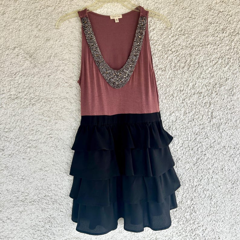Puede incluir: Vestido sin mangas con top malva y falda negra escalonada. La parte superior presenta un escote en V adornado con cuentas plateadas. El vestido est&aacute; colgado de una percha de pl&aacute;stico transparente sobre un fondo neutro.
