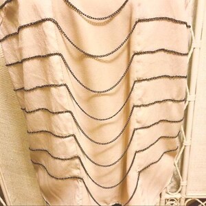 May include: A light peach-colored top with decorative black chain detailing. The chains are arranged in horizontal, curved lines across the front of the garment, creating a layered effect. The fabric appears to be a soft, flowing material.