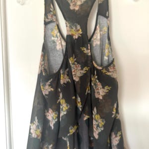 K&ouml;nnte beinhalten: Schwarzes Tanktop mit floralem Muster in Rosa, Gelb und Wei&szlig;. Das Oberteil hat ein Racerback-Design und besteht aus einem transparenten Stoff. Das Tanktop h&auml;ngt an einem durchsichtigen Kunststoffb&uuml;gel.
