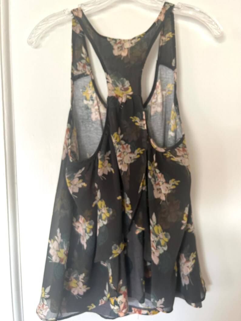 K&ouml;nnte beinhalten: Schwarzes Tanktop mit floralem Muster in Rosa, Gelb und Wei&szlig;. Das Oberteil hat ein Racerback-Design und besteht aus einem transparenten Stoff. Das Tanktop h&auml;ngt an einem durchsichtigen Kunststoffb&uuml;gel.
