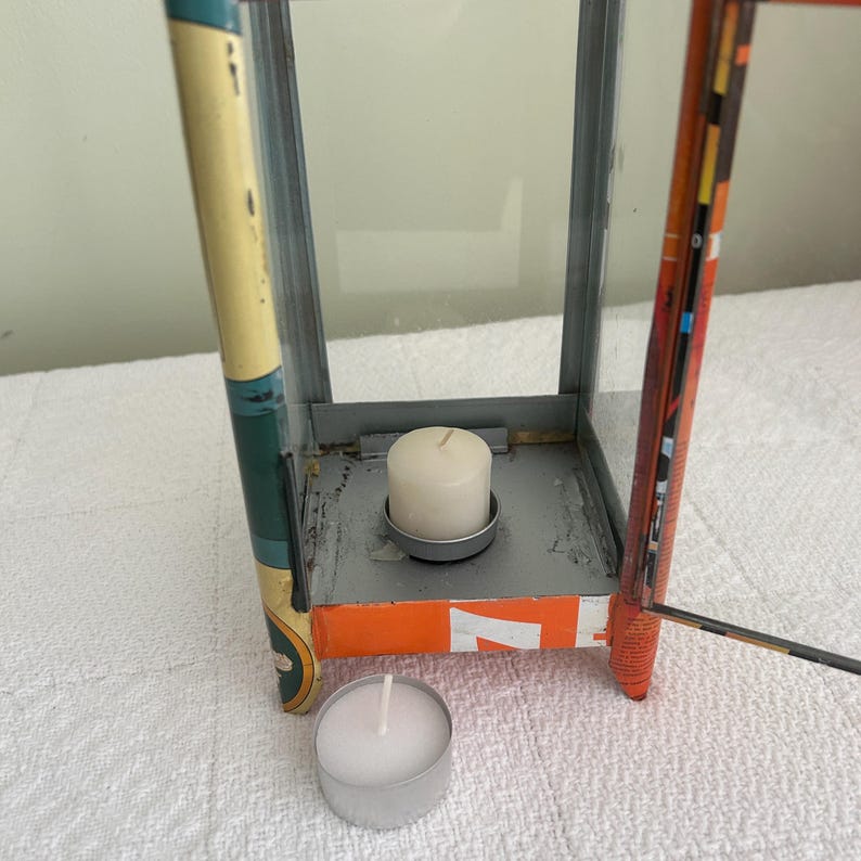 May include: A vintage metal lantern with glass panels, featuring a white candle inside and a tealight candle below. The lantern has orange and yellow accents, with a distressed finish, suggesting a rustic or industrial aesthetic.