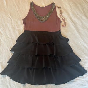 Puede incluir: Un vestido sin mangas con un corpi&ntilde;o malva y una falda negra escalonada. El escote est&aacute; adornado con cuentas plateadas. Un collar de cuentas rosas se coloca junto al vestido. El vestido est&aacute; sobre una superficie blanca.