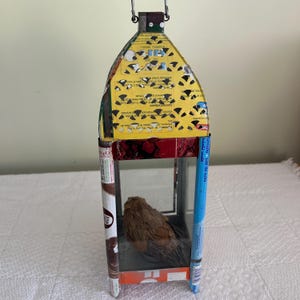 May include: A handmade lantern constructed from repurposed food packaging. The lantern features a yellow roof with cut-out designs, a clear glass enclosure, and a brown object inside. The frame is made from various colorful boxes and packaging materials.
