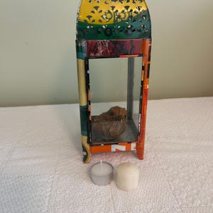 May include: A decorative metal lantern with a yellow, green, and orange design. Inside the lantern is a small, brown bird figurine. Two small candles sit in front of the lantern. The lantern is on a white surface.