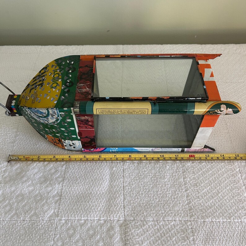 May include: A handmade, mixed-media birdhouse with a rectangular window and a cylindrical perch. The birdhouse is constructed from repurposed materials, including colorful paper and metal. The birdhouse measures approximately 14 inches long.