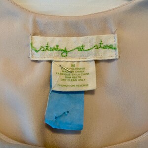 May include: Close-up of a garment tag with the text "story at stova" in green script. The tag also includes details about the material, size, and care instructions. A blue tag is attached to the garment tag.