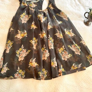 K&ouml;nnte beinhalten: Ein dunkelgraues Tanktop mit einem Blumenmuster in Gelb, Rosa und Wei&szlig;. Das Oberteil hat einen Rundhalsausschnitt und ein Racerback-Design. Der Stoff scheint leicht und halbtransparent zu sein, mit einer flie&szlig;enden Silhouette.