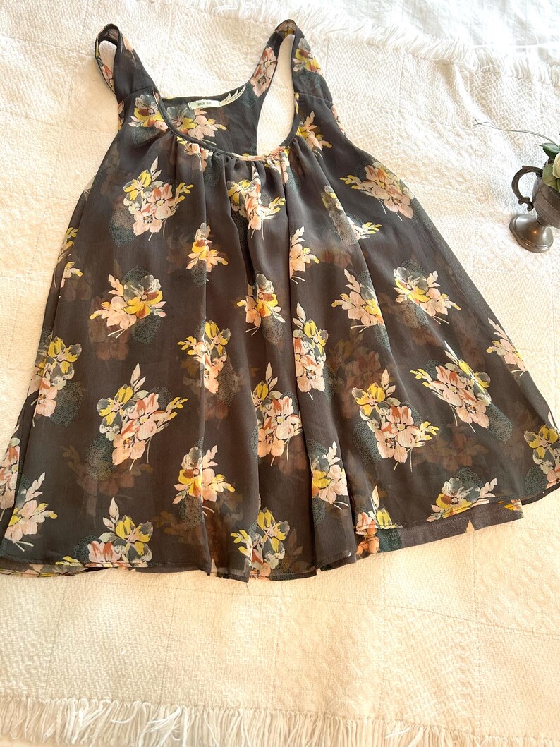 K&ouml;nnte beinhalten: Ein dunkelgraues Tanktop mit einem Blumenmuster in Gelb, Rosa und Wei&szlig;. Das Oberteil hat einen Rundhalsausschnitt und ein Racerback-Design. Der Stoff scheint leicht und halbtransparent zu sein, mit einer flie&szlig;enden Silhouette.