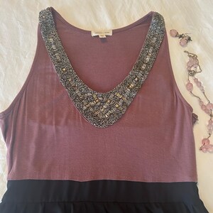 Puede incluir: Un vestido sin mangas de color malva con un escote con cuentas y una falda negra. Un par de pendientes rosas y un collar de cuentas rosas se colocan junto al vestido. El vestido tiene un escote en V.