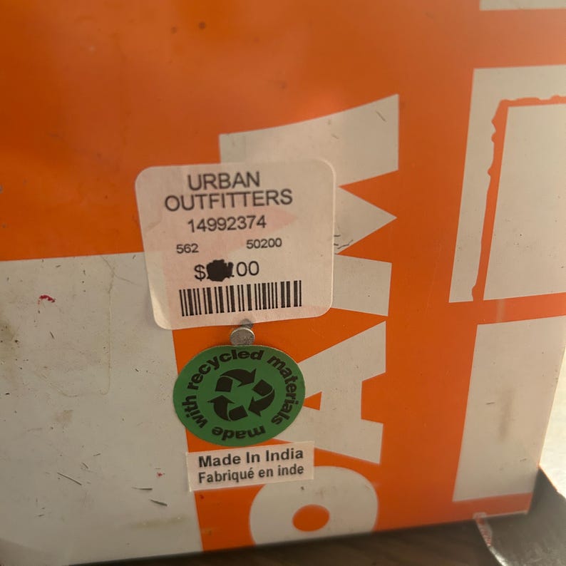 May include: Close-up of a white sticker with the text "URBAN OUTFITTERS" and product information. Below the sticker is a green circular sticker with a recycling symbol and the text "recycled materials". The background is orange.