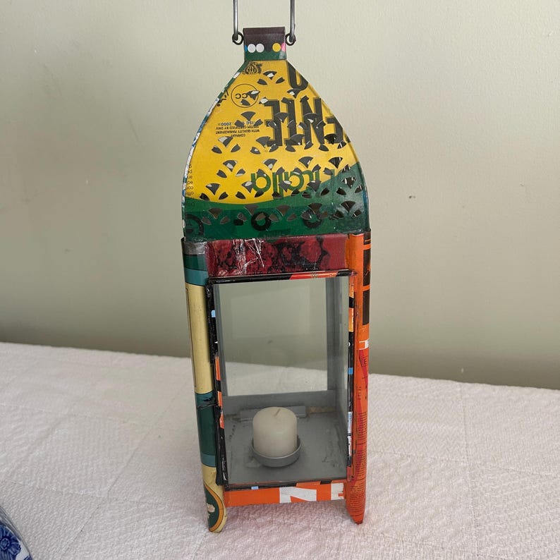 May include: A colorful metal lantern with a handle, featuring a rectangular glass window and a candle inside. The lantern is made from recycled materials, with a mix of orange, yellow, green, and red hues. The top has decorative cutouts.