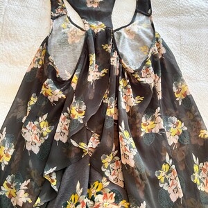 K&ouml;nnte beinhalten: Dunkelgraues Tanktop mit floralem Muster. Das Tanktop hat ein Racerback-Design und ist mit gelben, rosa und wei&szlig;en Blumen verziert. Der Stoff scheint leicht und flie&szlig;end zu sein, geeignet f&uuml;r warmes Wetter.
