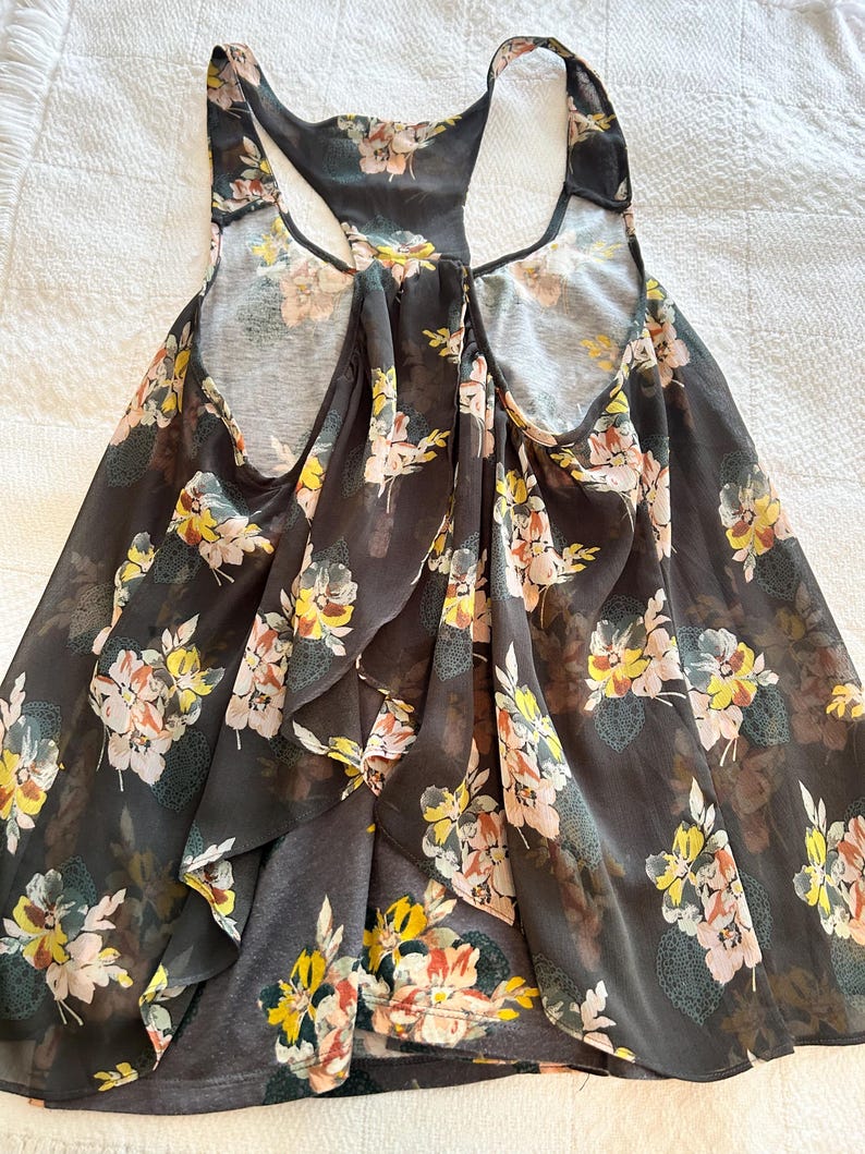 K&ouml;nnte beinhalten: Dunkelgraues Tanktop mit floralem Muster. Das Tanktop hat ein Racerback-Design und ist mit gelben, rosa und wei&szlig;en Blumen verziert. Der Stoff scheint leicht und flie&szlig;end zu sein, geeignet f&uuml;r warmes Wetter.