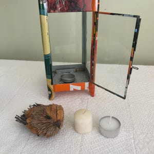 May include: A decorative metal lantern with an open glass door, orange and green accents, and a small candle holder inside. A decorative bird, a white pillar candle, and a tealight candle are next to the lantern.