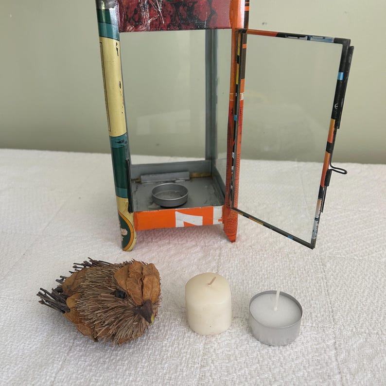 May include: A decorative metal lantern with an open glass door, orange and green accents, and a small candle holder inside. A decorative bird, a white pillar candle, and a tealight candle are next to the lantern.
