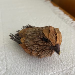 May include: A handcrafted bird sculpture made from natural materials. The bird features a body of dried grasses and leaves, with a beak and eye details. The sculpture is a warm brown color and rests on a white surface.