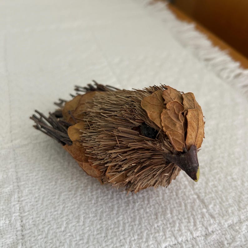 May include: A handcrafted bird sculpture made from natural materials. The bird features a body of dried grasses and leaves, with a beak and eye details. The sculpture is a warm brown color and rests on a white surface.
