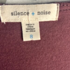 Puede incluir: Primer plano de una etiqueta de tela con la marca "silence + noise" en blanco. La etiqueta tambi&eacute;n dice "made in china, fabrique en chine" y el n&uacute;mero "8". El fondo es de color granate.