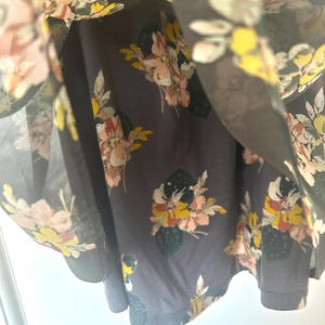K&ouml;nnte beinhalten: Nahaufnahme eines dunkelgrauen Kleides mit floralem Muster. Die Blumen sind in Rosa, Gelb und Wei&szlig; gehalten, mit gr&uuml;nen Bl&auml;ttern. Der Stoff wirkt leicht und flie&szlig;end.