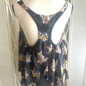 K&ouml;nnte beinhalten: Ein dunkelgraues Tanktop mit einem Blumenmuster in Rosa, Gelb und Wei&szlig;. Das Oberteil hat ein Racerback-Design und eine geschichtete, flie&szlig;ende Silhouette. Der Stoff wirkt transparent und leicht.