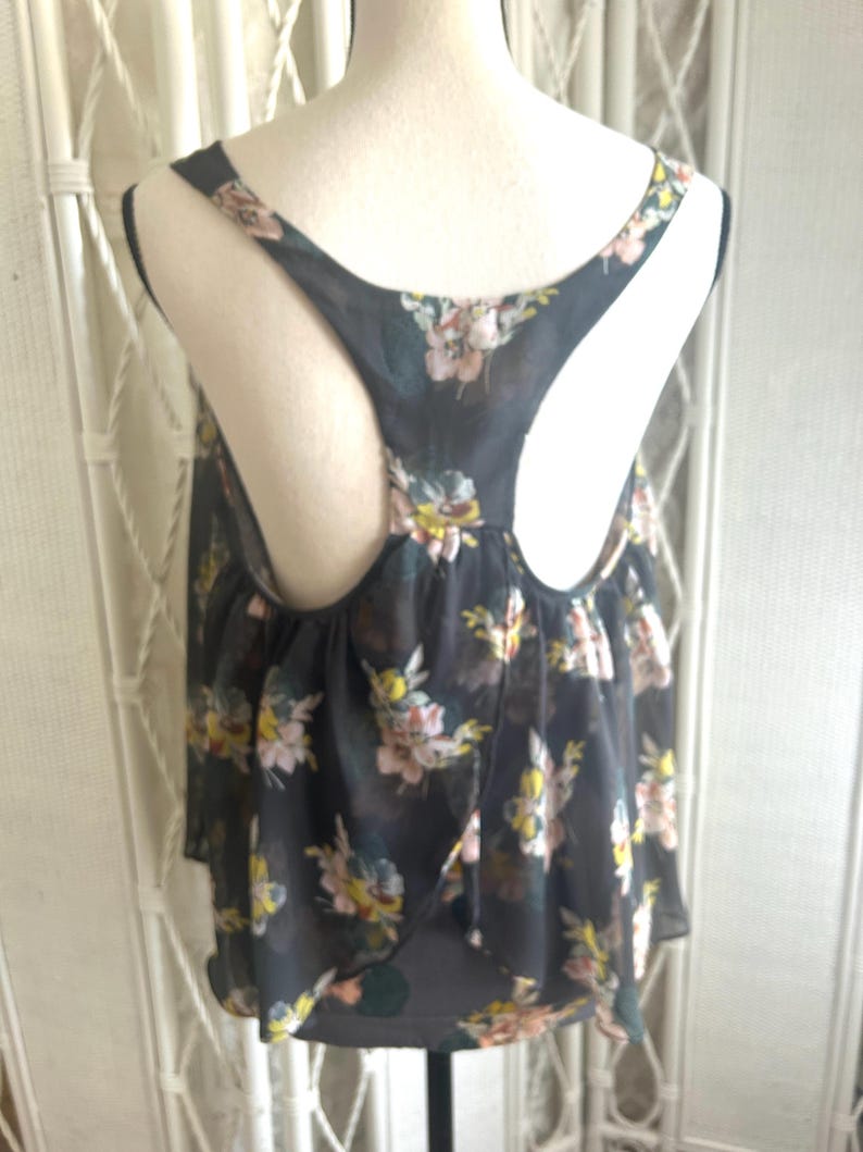 K&ouml;nnte beinhalten: Ein dunkelgraues Tanktop mit einem Blumenmuster in Rosa, Gelb und Wei&szlig;. Das Oberteil hat ein Racerback-Design und eine geschichtete, flie&szlig;ende Silhouette. Der Stoff wirkt transparent und leicht.