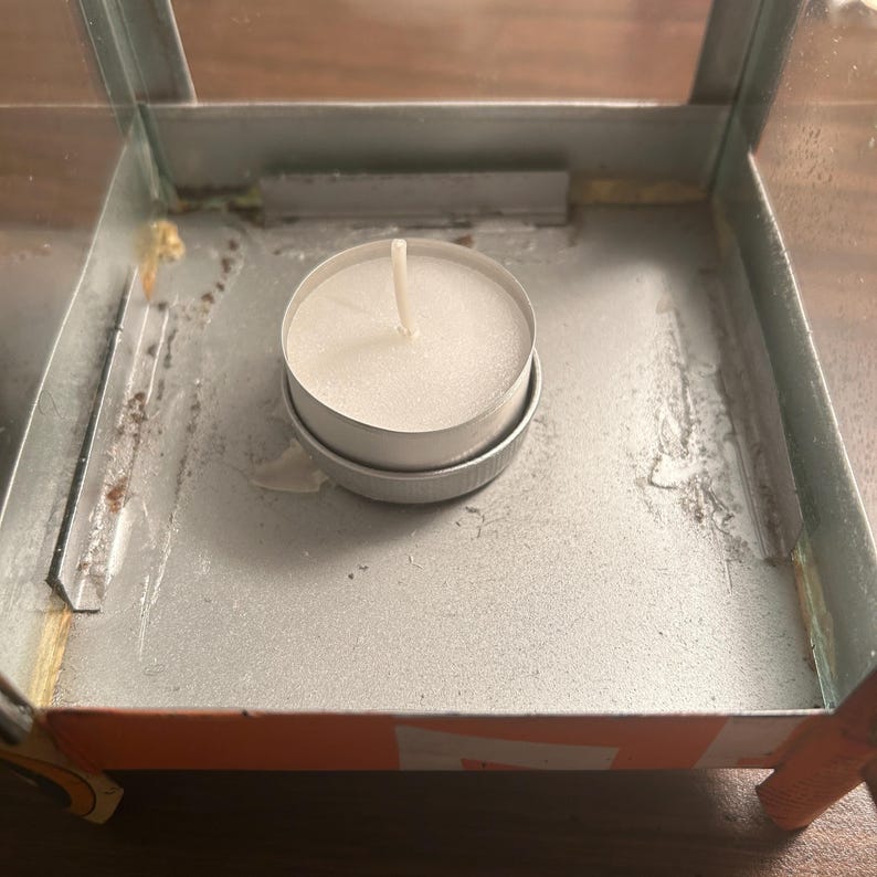 May include: A small, round white candle in a silver metal container sits inside a rectangular metal lantern. The lantern has glass sides and a metal frame. The candle has a white wick.