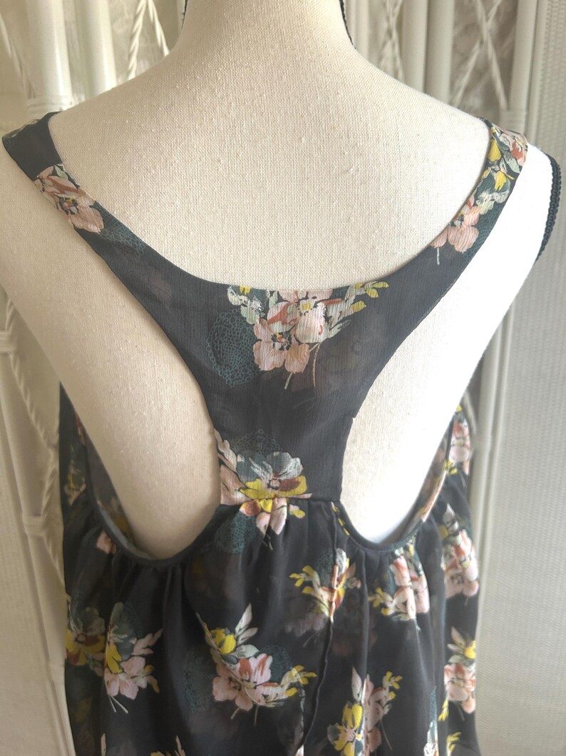 K&ouml;nnte beinhalten: Schwarzes Tanktop mit einem Blumenmuster in Rosa, Gelb und Gr&uuml;n. Das Oberteil hat ein Racerback-Design und besteht aus einem transparenten Stoff. Der Blumendruck ist &uuml;ber den Stoff verstreut.