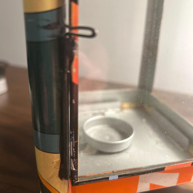 May include: A vintage metal lantern with a clear glass enclosure. The lantern has a small, round metal base inside and a handle on top. The exterior features orange, blue, and yellow painted details. The lantern is designed to hold a small candle.