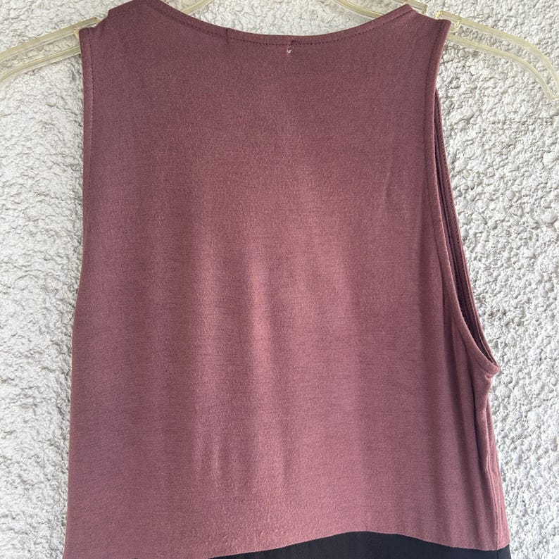 Puede incluir: Una camiseta sin mangas, con cuello redondo, de color malva apagado, que pasa a negro en el dobladillo. La tela parece suave y se adapta suavemente. La camiseta est&aacute; colgada de una percha de pl&aacute;stico transparente sobre un fondo texturizado y claro.